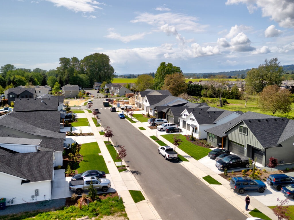 Aerial view of a residential neighborhood with houses, driveways, cars, and green fields under a partly cloudy sky